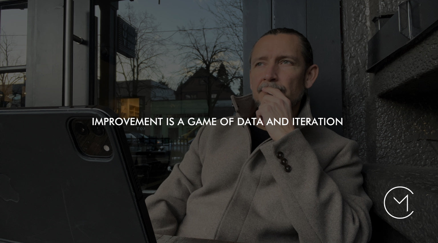 A man sits thoughtfully with a tablet at an outdoor table, illustrating reflection, data and iteration in a trading journal.